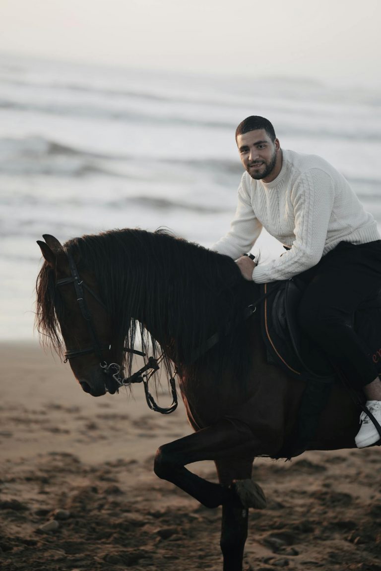 A man in a white sweater rides a horse on Essaouira beach, Morocco, during sunset.