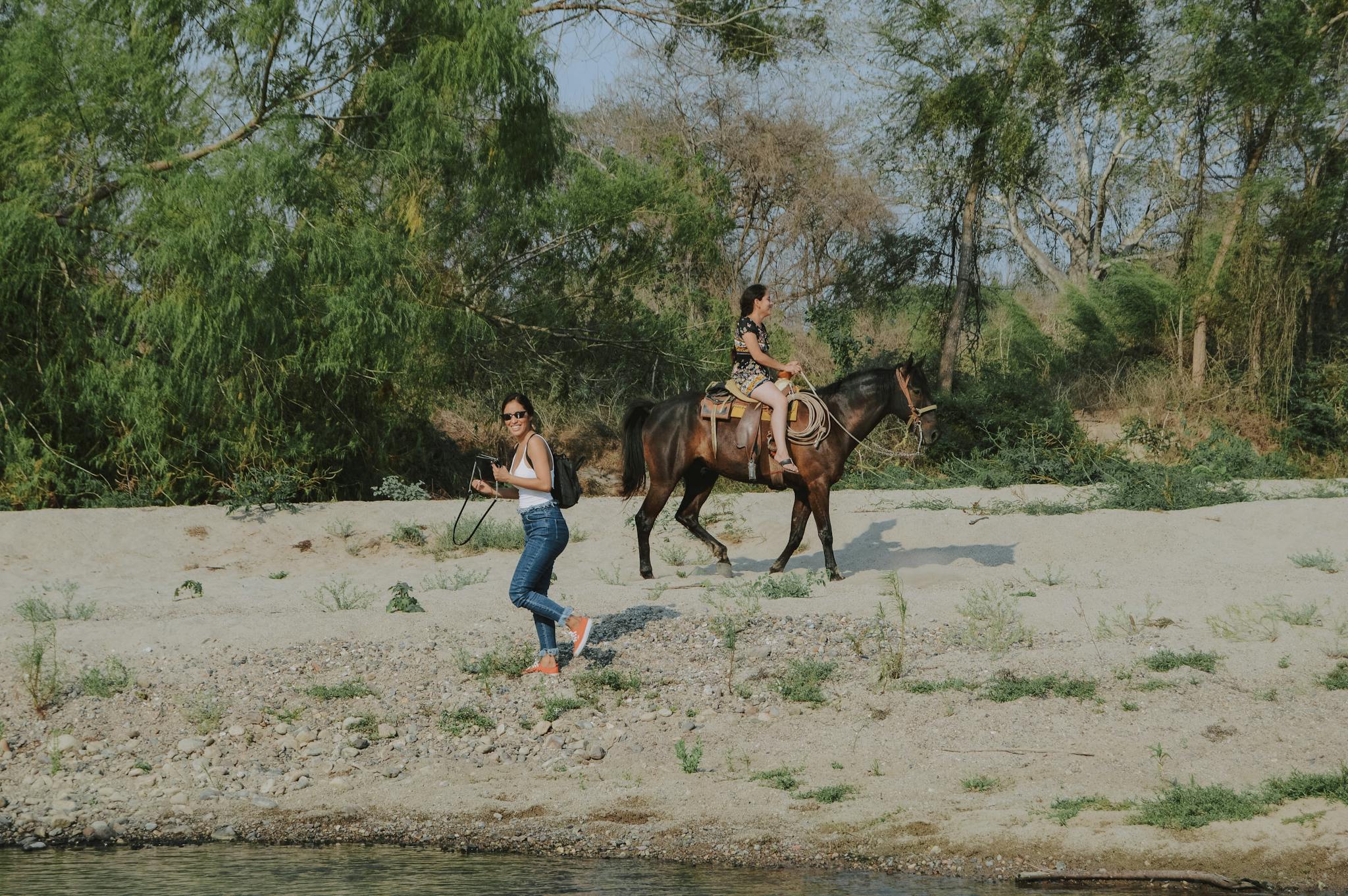 Two women enjoying a horseback ride by a river, surrounded by lush greenery.