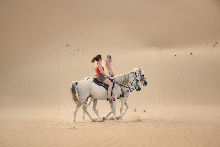 Two women riding horses through the dunes of Dubai's desert, capturing serene beauty and adventure.